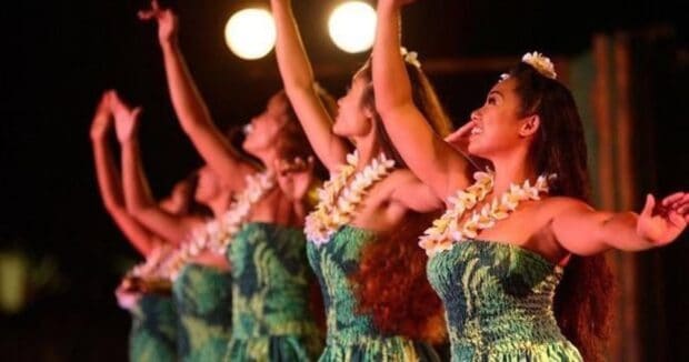 Four Germaine's Luau hula dancers in green print dresses and plumeria lei with arms raised on the night stage