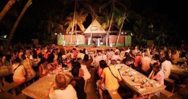 Guests eating at long picnic tables on the sand facing the Germaine's Luau stage at night