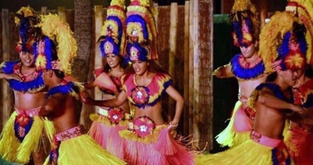 Germaine's Luau dancers in feather headdresses and grass skirts mid-Tahitian dance in front of a wood fence