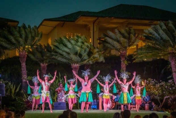 Grand Hyatt Kauaʻi lūʻau dancers in Tahitian costume on the grass stage, with a resort building and palms behind them