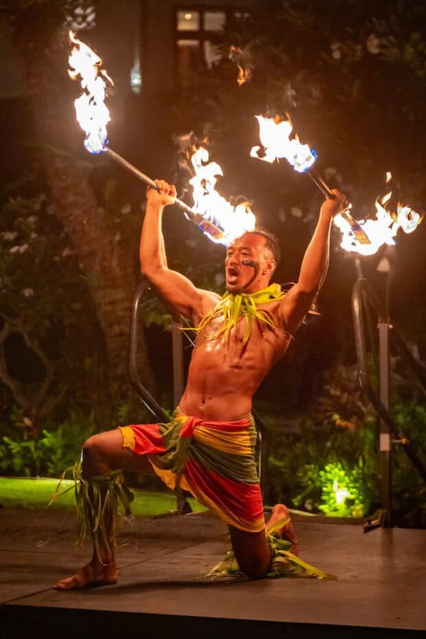 Fire-knife dancer kneeling with two flaming knives raised overhead at the Grand Hyatt Kauaʻi lūʻau