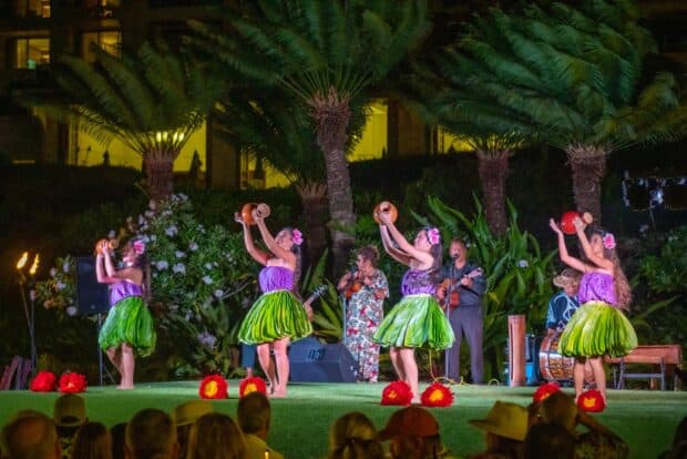 Hula dancers in purple tops and green skirts raise their arms on the Grand Hyatt Kauaʻi lūʻau stage