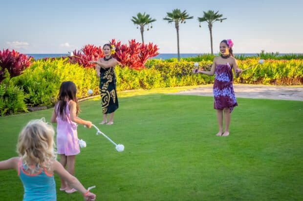 Two dancers teach children to twirl poi balls on a lawn at the Grand Hyatt Kauaʻi lūʻau