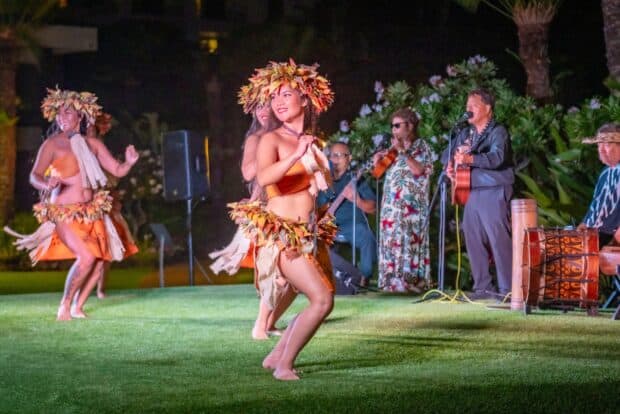 Dancer in a feathered headpiece and orange Tahitian costume smiles mid-dance, with musicians behind her