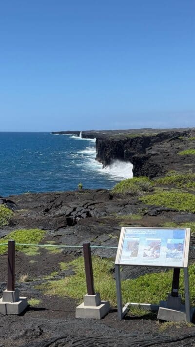 Lava sea cliffs and surf from the Hōlei Sea Arch viewpoint, with a park sign about the changing coastline