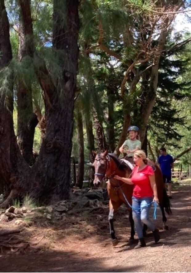 Guide leading a pony ride with a young child in a helmet under tall trees at Ironwood Ranch