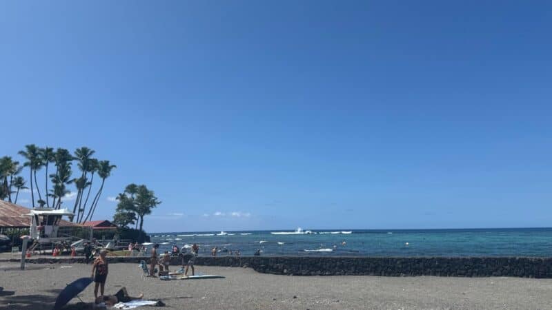 Dark sand beach at Kahaluʻu Beach Park with the lava-rock seawall, calm inner bay and waves breaking on the outer reef