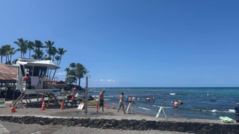 Lifeguard tower with orange cones at Kahaluʻu Beach Park, people walking along the seawall and snorkelers in the bay