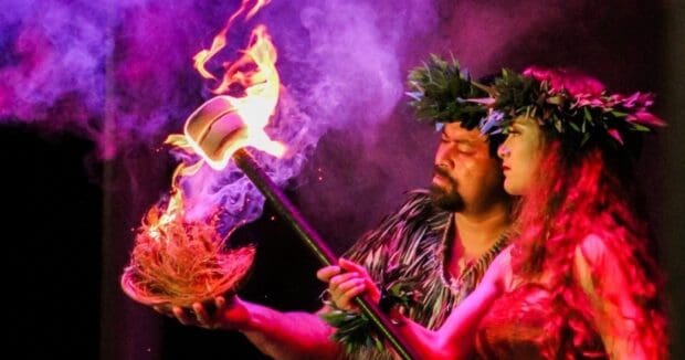 Two performers holding a burning coconut-husk bowl and torch in purple stage light at Ka Moana Luau