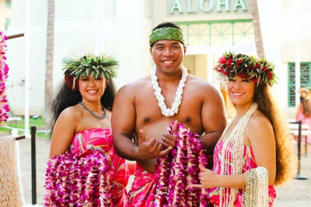 Three Ka Moana Luau greeters holding purple orchid lei below the ALOHA lettering at the entrance