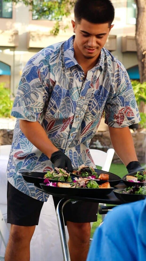 Server in an aloha shirt delivering plated salad courses from a tray stand