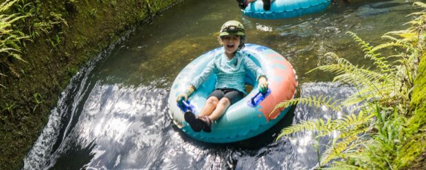 A young child in a helmet and gloves laughs while floating an irrigation canal on the Kauaʻi mountain tubing tour