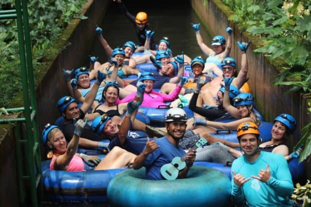 A large tubing group in helmets and headlamps fills a plantation flume, a guide in front holding a ukulele