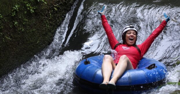 A tuber in a helmet and gloves rides over a small drop in a mossy irrigation canal on the Kauaʻi tubing tour