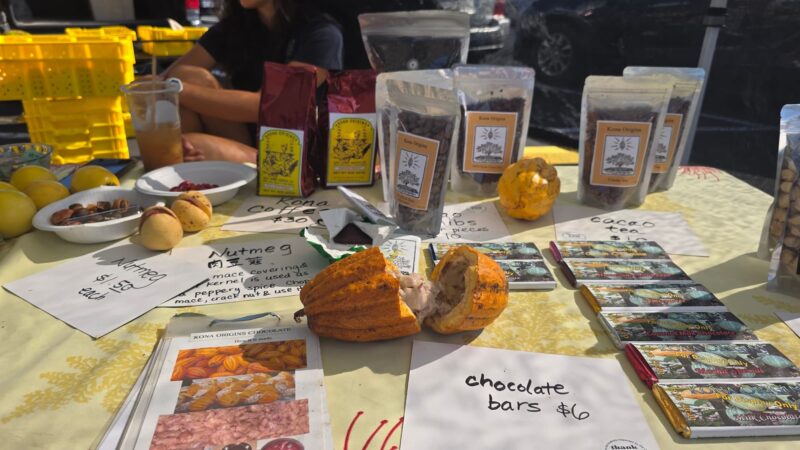 Kona Origins table with Kona coffee, a split cacao pod, chocolate bars, and nutmeg at the Keauhou Farmers Market