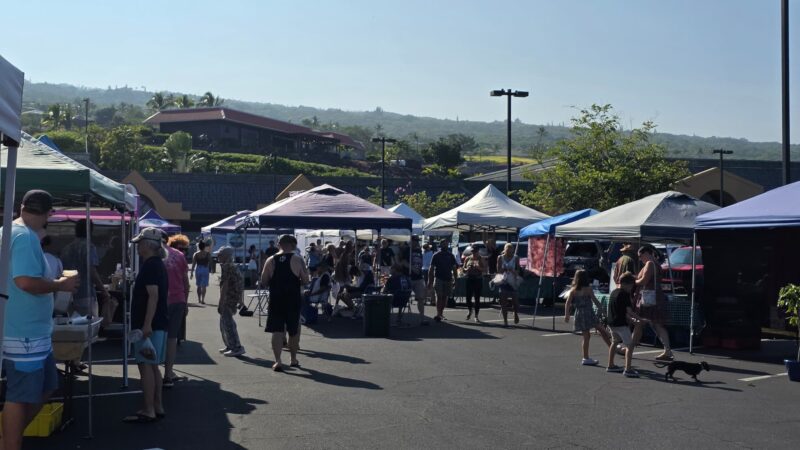 Shoppers walking between vendor tents at the Keauhou Farmers Market with green hills behind