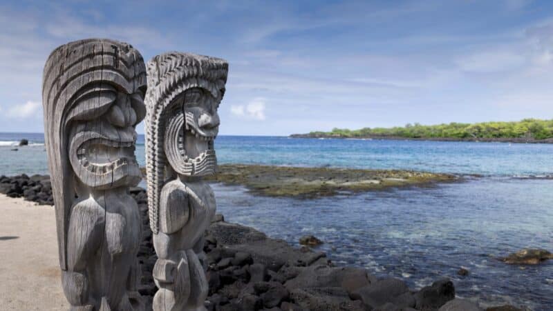 Two carved wooden kiʻi at the water’s edge at Puʻuhonua o Hōnaunau, facing a calm cove with a low lava shelf