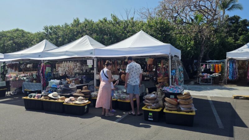 Two shoppers browsing sun hats and woven beach bags at a Kona Farmers Market craft stall