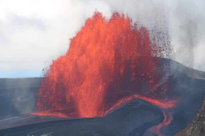 Tall orange lava fountain erupting from a vent cone, with lava streaming across a dark crater floor