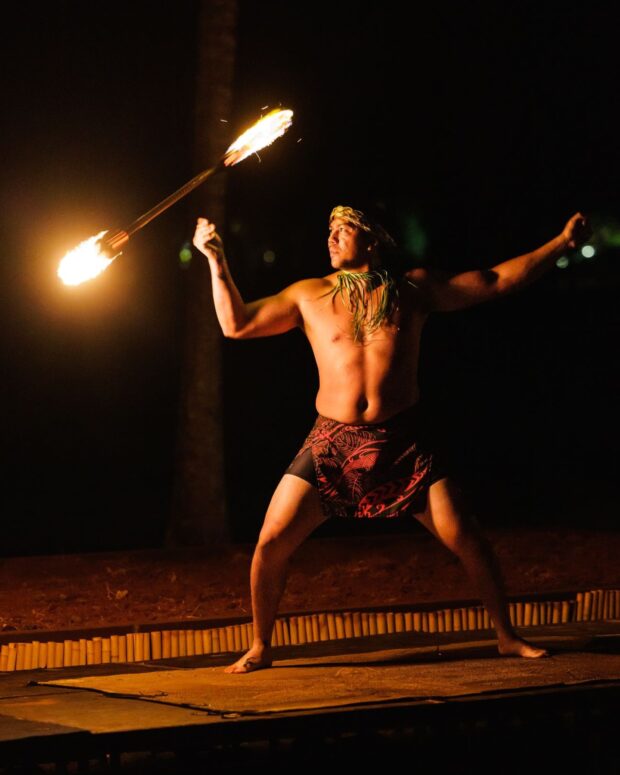 Fire-knife dancer holding a knife lit at both ends on a low stage at night, Lūʻau Ka Hikina