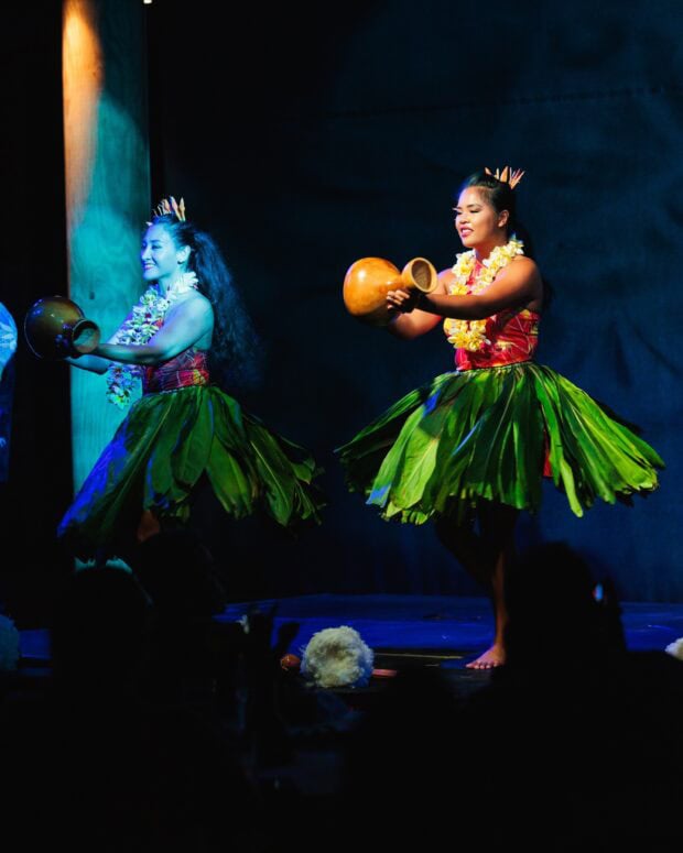 Two hula dancers in green ti-leaf skirts holding ipu gourds on the Lūʻau Ka Hikina stage
