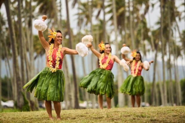 Three hula dancers shaking feathered gourd rattles on a lawn under coconut palms, Lūʻau Ka Hikina