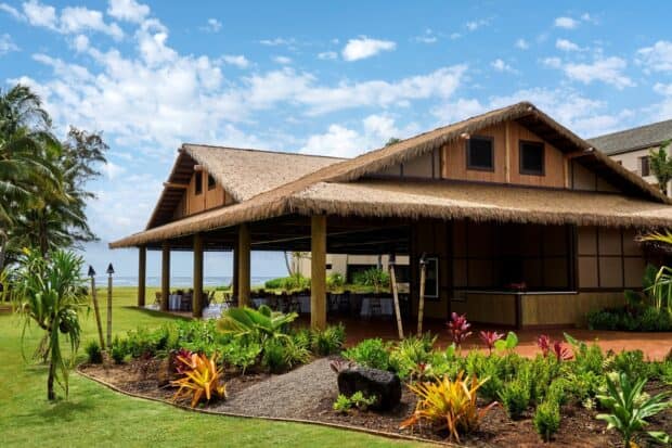 Thatched open-air Hālau Hoʻokipa pavilion on a lawn with the ocean behind it, the Lūʻau Ka Hikina venue in Kapaʻa
