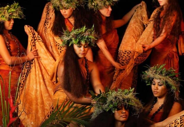 Women dancers in orange with fern crowns and patterned cloth wraps kneel and stand in a Luau Kalamaku story scene