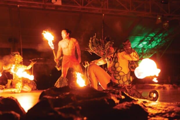 Fire dancers with torches and a costumed performer on the rock-edged Luau Kalamaku stage under the pavilion's steel roof