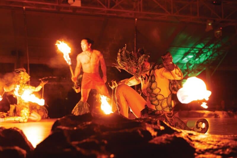 Fire dancers with torches and a costumed performer on the rock-edged Luau Kalamaku stage under the pavilion's steel roof