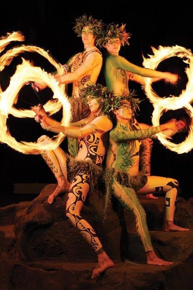 Four body-painted performers with fern crowns spin flaming fire poi on stage rocks at Luau Kalamaku