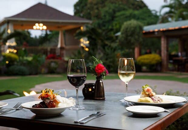 Plated dishes and glasses of red and white wine on a courtyard table at Gaylord's at Kilohana, lawn and gazebo behind