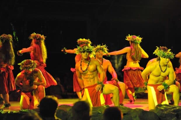 Hula dancers in red with leaf crowns and lei on the Luau Kalamaku stage, audience heads in the foreground
