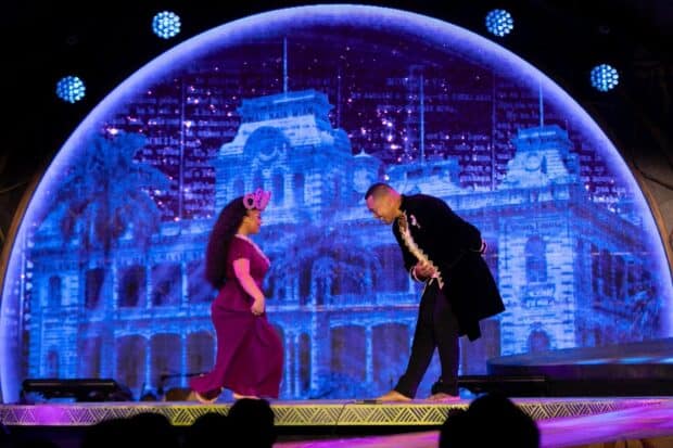 Performers in a crimson gown and a black frock coat on stage before a projection of ʻIolani Palace, Kaula Luau, Ko Olina