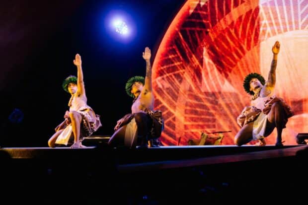 Three male dancers kneel with right arms raised, in leaf head lei and kapa-print wraps, at the Kaula Luau in Ko Olina