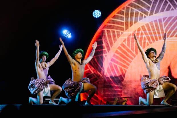Three male dancers kneel with both arms raised against an orange projected backdrop at the Kaula Luau, Ko Olina