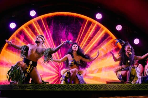 Three dancers kneel in yellow and black kapa-print costumes under pink stage lights at the Kaula Luau in Ko Olina