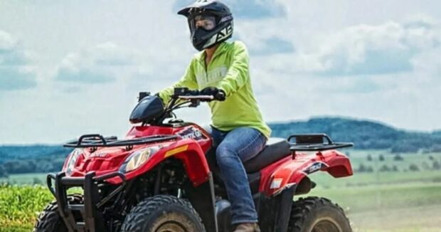 Rider in a helmet and goggles seated on a red four-wheel ATV with open pasture and hills behind
