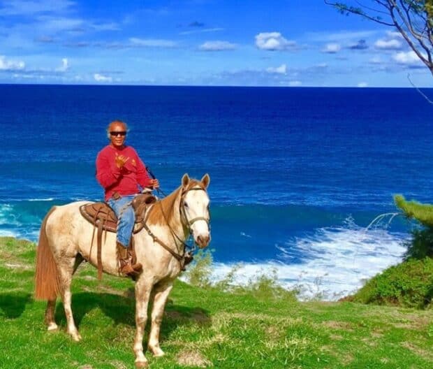 Smiling rider in a red top giving a shaka on a pale horse on a grassy bluff above the ocean, Maui horseback ride