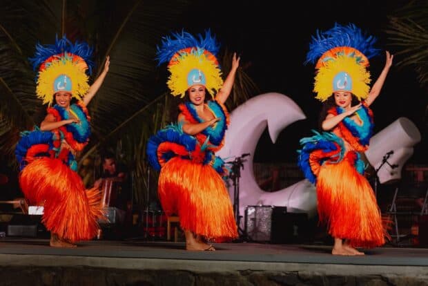Three dancers in tall feather headdresses and orange grass skirts on stage at the Mauka Warriors Luau in Kapolei