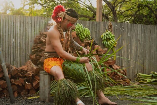 A performer weaves a green coconut frond beside stacked firewood and green banana bunches at the Mauka Warriors Luau, Kapolei