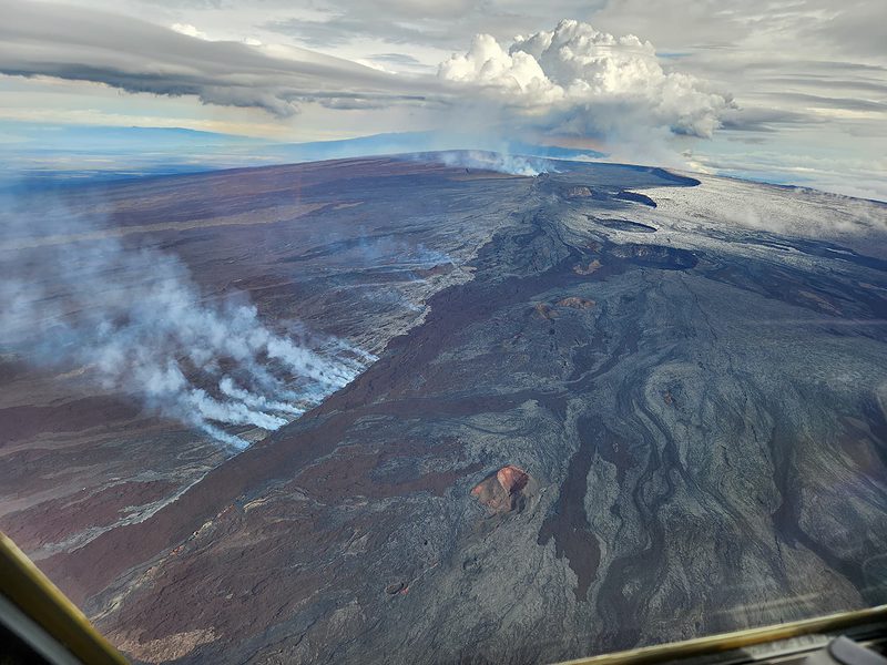 Aerial view of Mauna Loa summit with Lua Hou in the foreground followed by Lua Hohonu, South Pit, and Mokuʻāweoweo summit caldera. A small dusting of snow covers the ground east of the summit. The steaming fissure was active during the beginning of the eruption, but this morning the lava in the summit caldera had already cooled to a black color. The white plume in the background is rising from the fissures in the Northeast Rift Zone. USGS photo J. Schmith