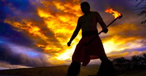 Fire dancer holding a lit fire knife silhouetted against a sunset sky at Huakaʻi Lūʻau, Maui