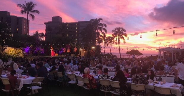 Guests dining at long tables on the lawn as a hula dancer performs at sunset, Huakaʻi Lūʻau, Kāʻanapali, Maui