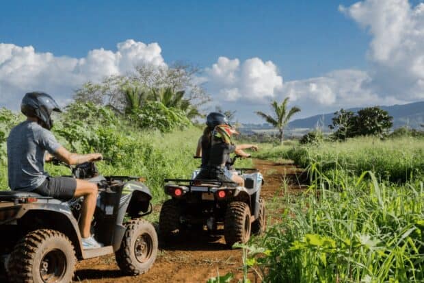 Two riders on their own ATVs on a dirt farm trail with the coast and green fields behind