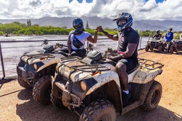 A single rider on a mud-splashed ATV on a dirt trail lined with green foliage