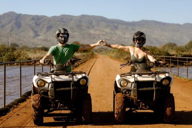 Two riders on ATVs making a heart shape with their hands on a dirt causeway with mountains behind