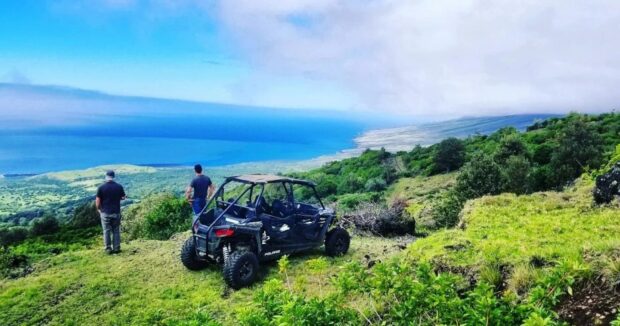 Two people standing beside a side-by-side ATV looking out over the West Maui coastline from a green ridge