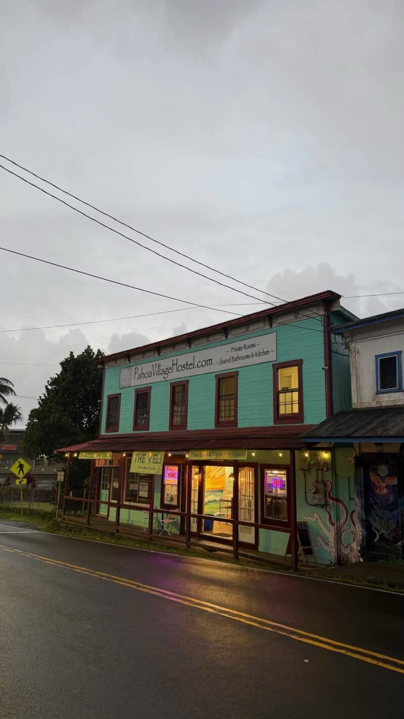 Teal two-story Pāhoa Village Hostel on the main street in Pāhoa at dusk, wet road in the foreground