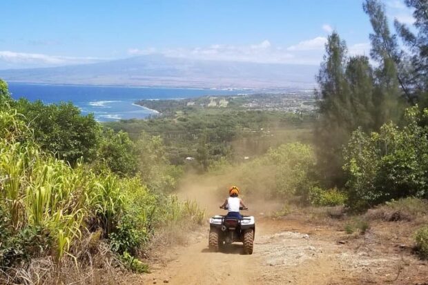 ATV rider heading down a dirt road with the West Maui coastline, ocean, and Haleakala in the distance